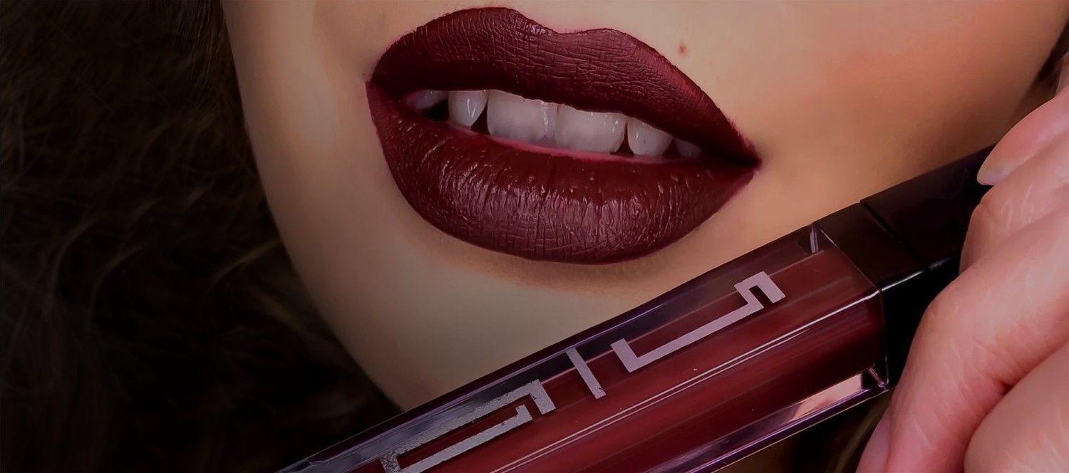 Close-up of lips wearing a bold, deep burgundy matte lipstick. The person holds the matching Timeless Beauty lipstick tube, showcasing its rich color and sleek packaging.